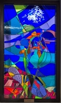 Abstract stained-glass panel in a dark wood frame featuring swirling shades of deep blue, turquoise, and violet. At the center, a cluster of bright orange, red, and green glass forms an energetic focal point surrounded by curved blue shapes suggestive of waves or motion. A white splash-like form appears near the top against a vivid cobalt-blue background. The lower section contains fragmented red, pink, purple, and yellow pieces intersected by dark lead lines, creating a sense of tension and movement. by Stephen Wilson